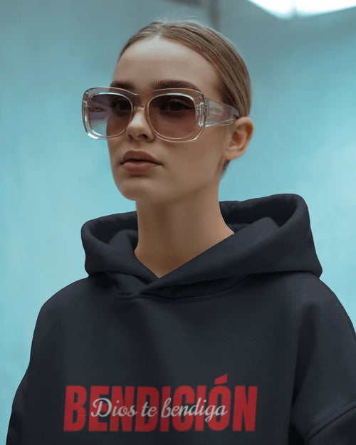 person wearing a navy bendicion unisex hoodie displayed on a light blue background with bold red bendición text across the chest and smaller white script layered over the center of the design designed by iron and velvet 62