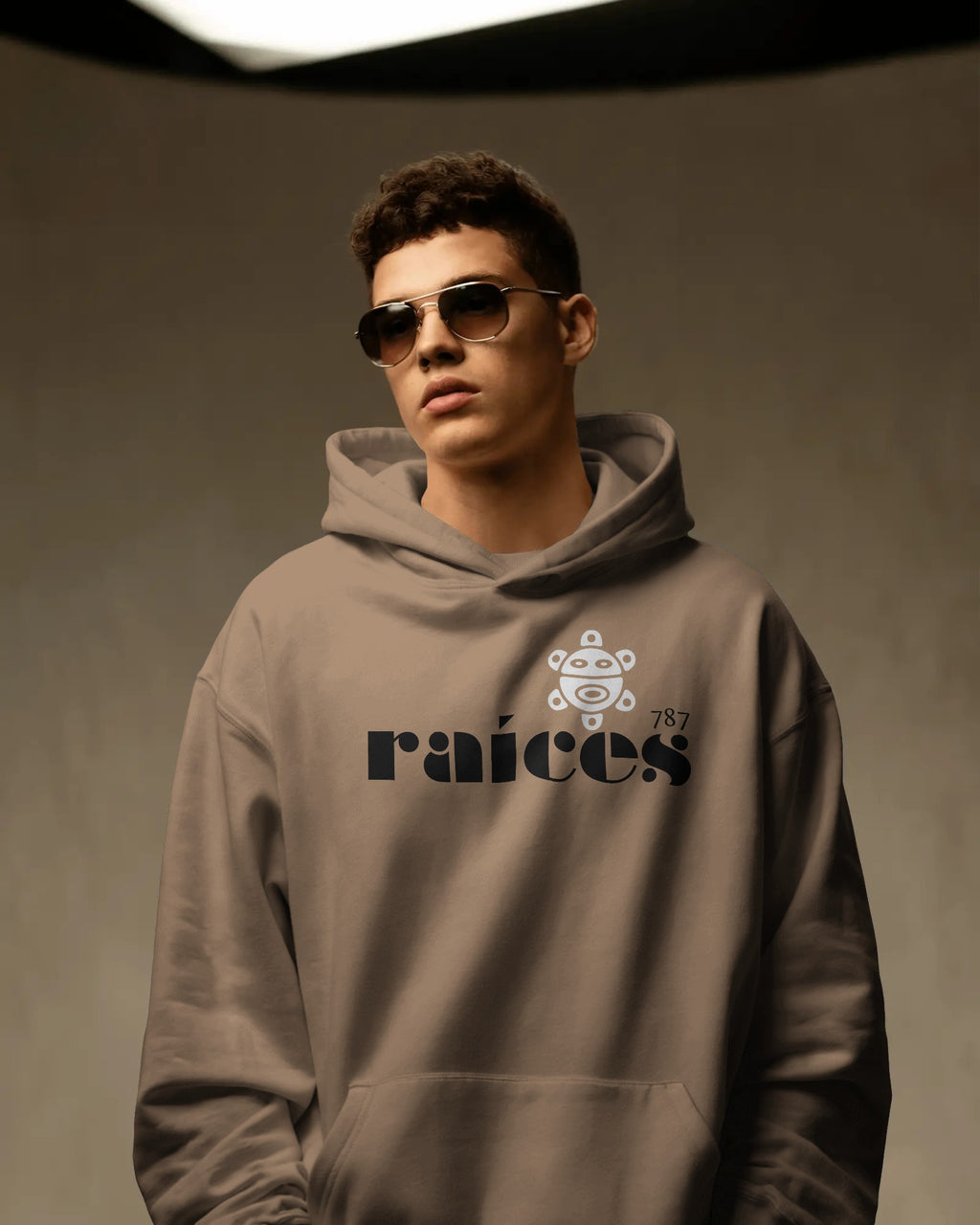 person in latte color unisex hoodie displayed on a neutral background with bold black raíces text across the chest, a white taíno sun and 787 detail, puerto rican cultural design by iron and velvet 62