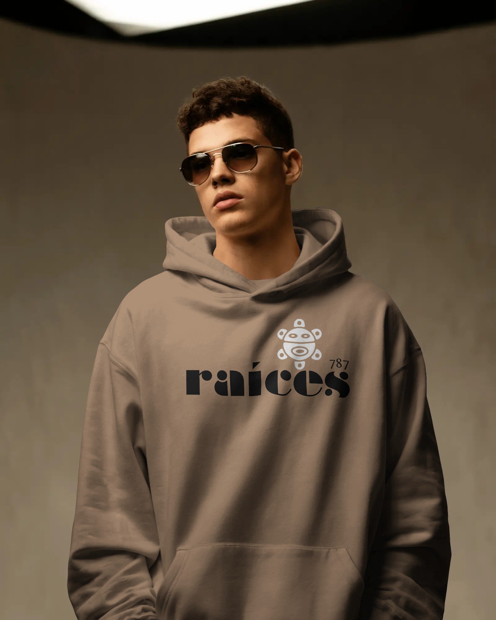 person in latte color unisex hoodie displayed on a neutral background with bold black raíces text across the chest, a white taíno sun and 787 detail, puerto rican cultural design by iron and velvet 62